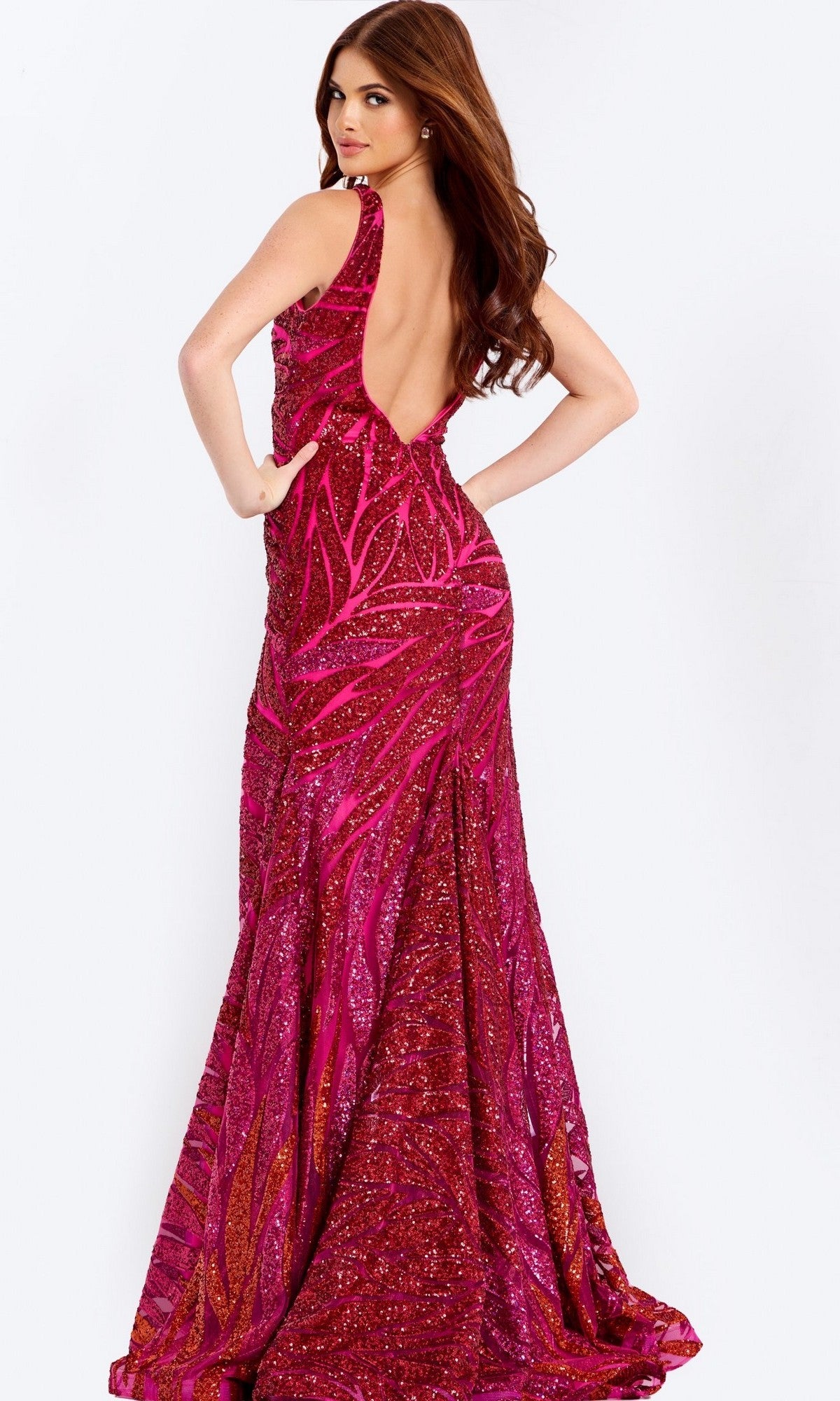 A woman stands facing away, looking over her shoulder, wearing the Jovani 47682 low-back long sequin prom dress in sparkling pink and red.
